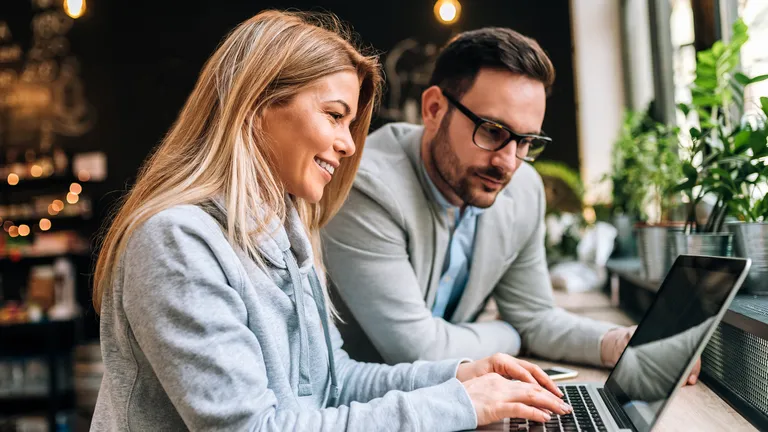 Eine Frau und ein Mann stehen nebeneinander in einem Café an einem Stehtisch und schauen auf einen Laptop.