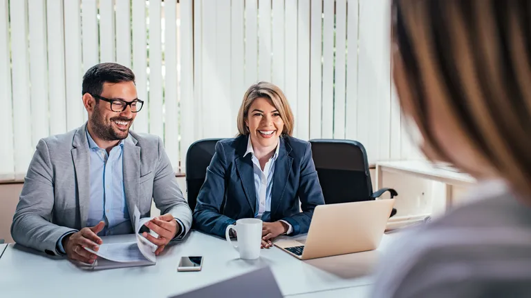 Woman and man in suits sitting at a table in an office looking at another woman.