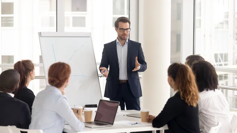 Six people sit at a table, a man stands in front of them next to a flipchart and looks at them, gesticulating.