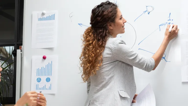 Woman with long wavy hair stands at the whiteboard and writes something on it. A man and a second woman are sitting at a table in front of it and watching.