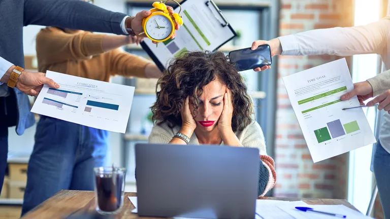 Woman sits in front of her laptop and rests her head on her hands. People are standing around her and holding things out to her.