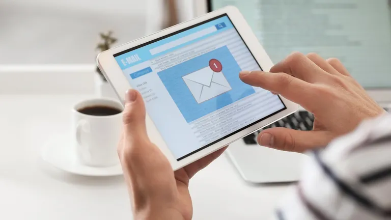 Someone holds a tablet in their left hand and taps the e-mail icon on the tablet with their right hand.