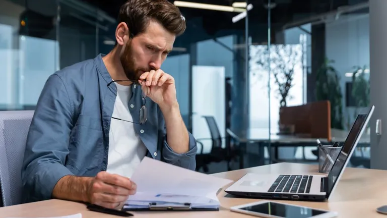 A man sits in front of a laptop and frowns at a sheet of paper he is holding in his hand.