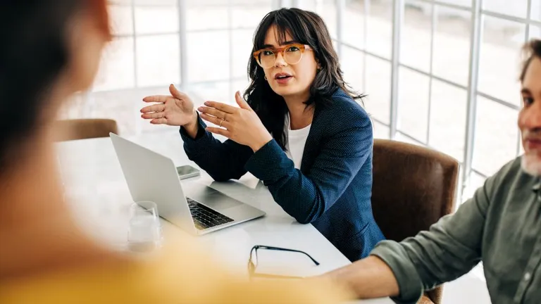 A woman and a man are sitting at a meeting table with a laptop in front of the woman, she is looking at a third person, of whom only the back of her head and shoulders are visible.