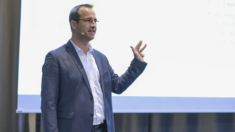 Speaker from the 2023 keynote event in a suit and with a microphone stands in front of a screen and sticks up four fingers.