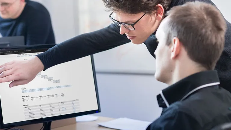 Mann sitzt vor Computerbildschirm ein anderer Mann steht neben ihm und zeigt auf etwas auf dem Bildschirm.