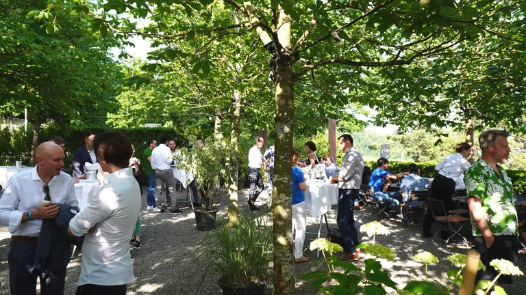 Gravel square with trees, people stand on the square at bar tables and talk to each other.