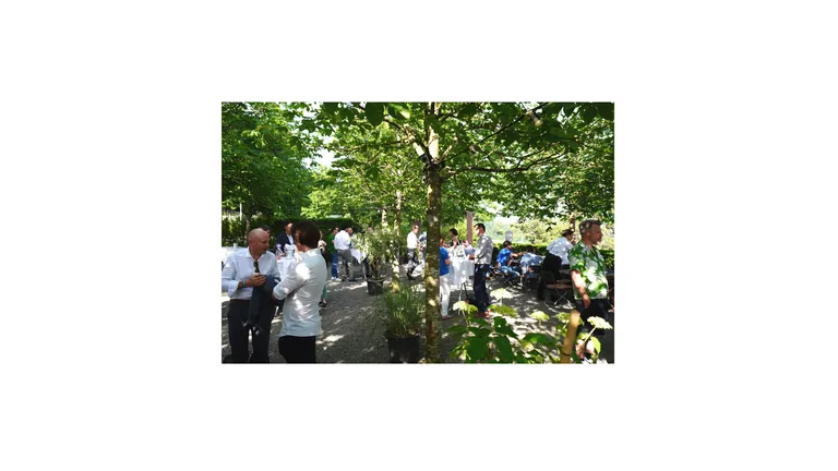 Gravel square with trees, people stand on the square at bar tables and talk to each other.
