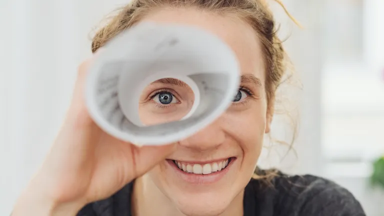 Young woman holds a rolled-up sheet of paper in front of her eye and looks through it.