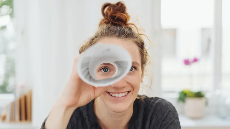 Young woman holds a rolled-up sheet of paper in front of her eye and looks through it.