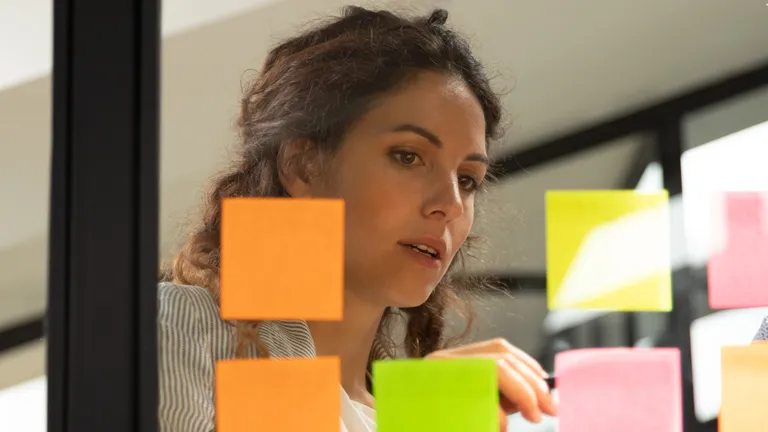 Woman and man stand in front of a glass wall with sticky notes and look at one of the notes.