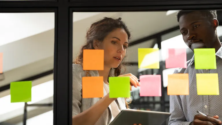 Woman and man stand in front of a glass wall with sticky notes and look at one of the notes.
