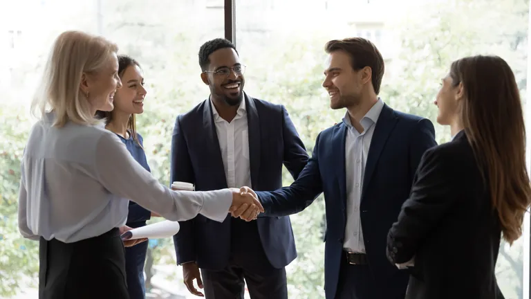 Three women and two men, all in suits, stand in a circle. A man and a woman shake hands.