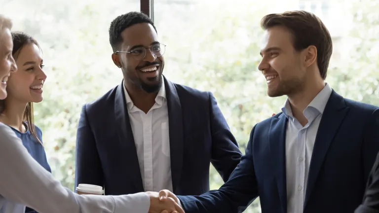 Three women and two men, all in suits, stand in a circle. A man and a woman shake hands.