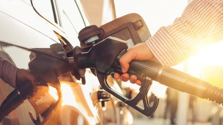 A hand holds a petrol hose and fills up a black car. The golden evening sun can be seen in the background.